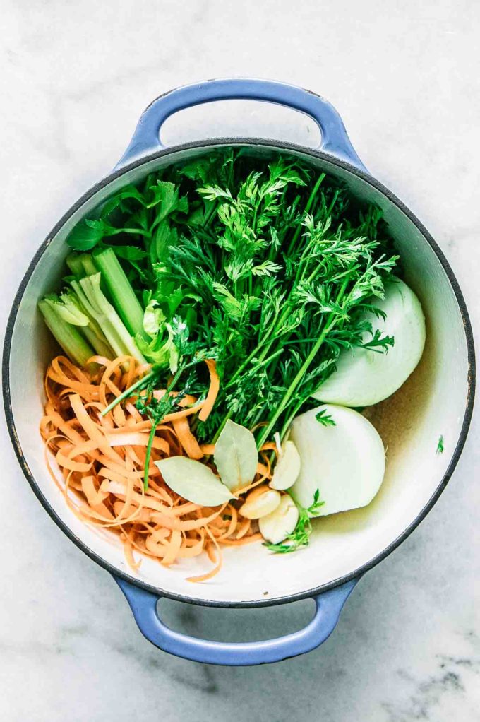 carrot leaves, carrot peels, onion, garlic, and celery in a large soup pot on a white table