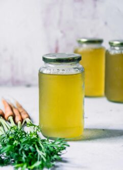 a glass jar of vegetable broth made from carrot tops on a white table with carrots with green leaves in the foreground