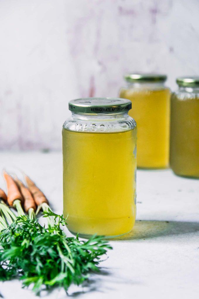 a glass jar of vegetable broth made from carrot tops on a white table with carrots with green leaves in the foreground