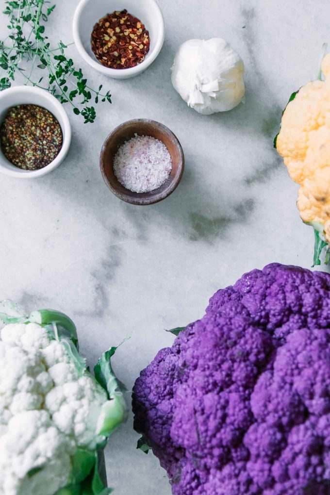white, purple, and yellow heads of cauliflower on a white table with three small bowls filled with salt, mustard, and red pepper flakes
