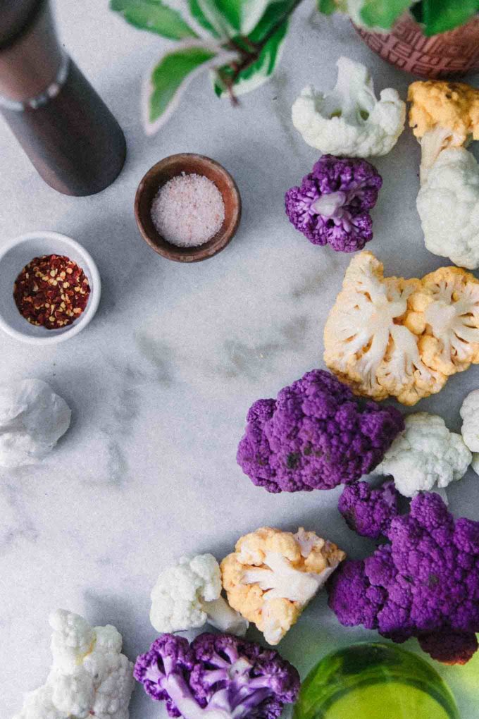purple, white, and orange cauliflower on a white table with a bowl of salt and red pepper