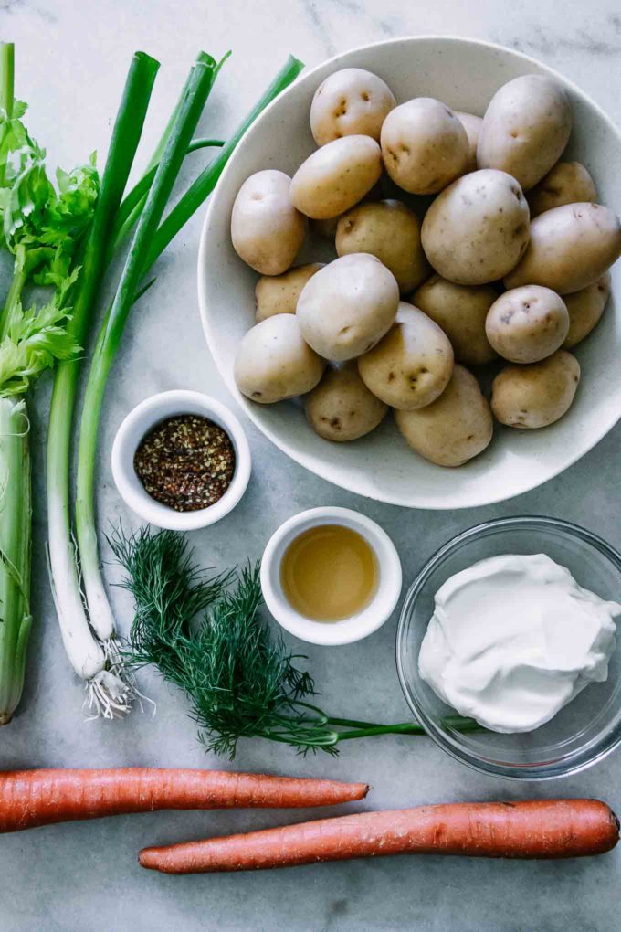 ingredients for a potato salad on a white table, including potatoes, green onions, celery, carrots, dill, vinegar, mustard, and vegan mayonnaise