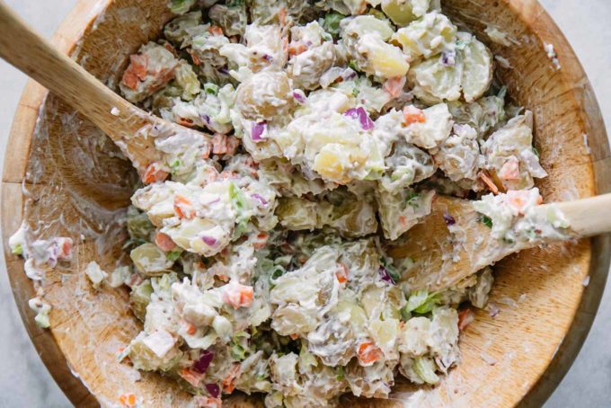 a closeup of completed potato salad in a large wooden bowl with two wooden spoons