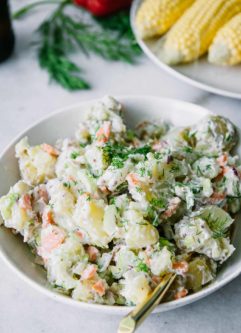vegan potato salad with mustard and dill in a white bowl on a table with picnic vegetables