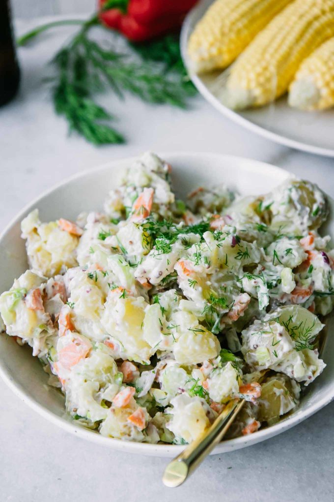 vegan potato salad with mustard and dill in a white bowl on a table with picnic vegetables