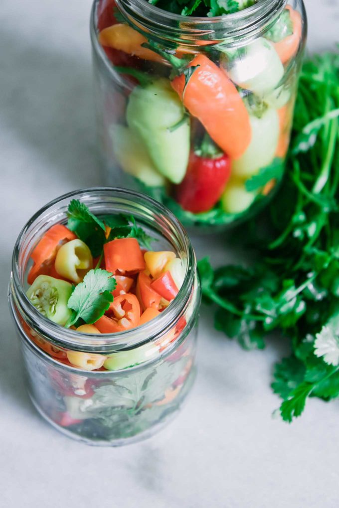 sliced and whole peppers in glass jars with fresh herbs and spices before pickling