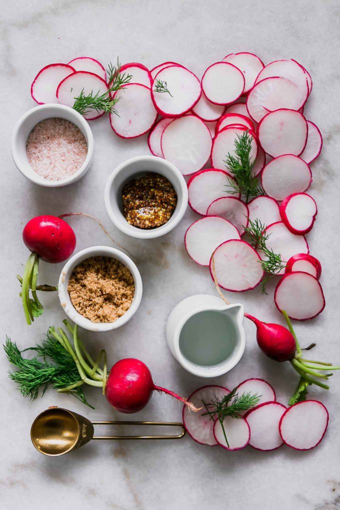 sliced radishes and small bowls of sugar, salt, vinegar, water, and fresh dill on a white table