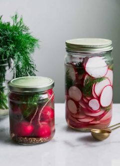 two jars of sliced and whole pickled radishes on a white table with fresh dill