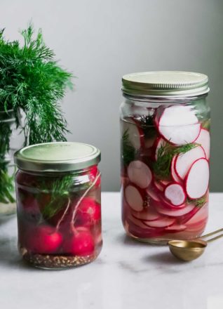 two jars of sliced and whole pickled radishes on a white table with fresh dill