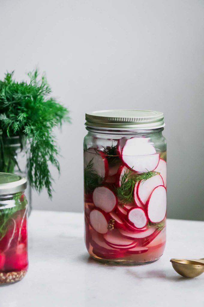 a jar of sliced pickled radishes on a white table with fresh dill