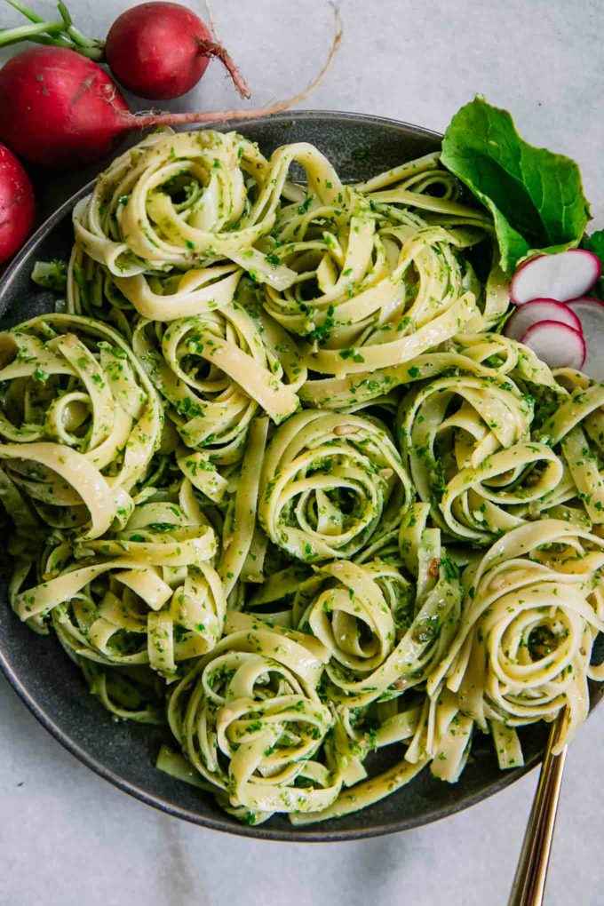 a close up of pesto fettuccine on a dark plate with a gold fork and red radishes in the background