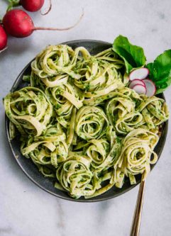 pesto pasta on a blue plate with a gold fork on a white table with red radish