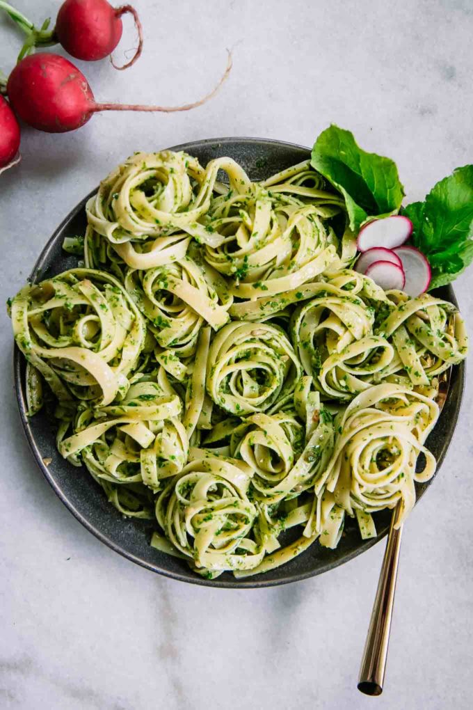pesto pasta on a blue plate with a gold fork on a white table with red radish