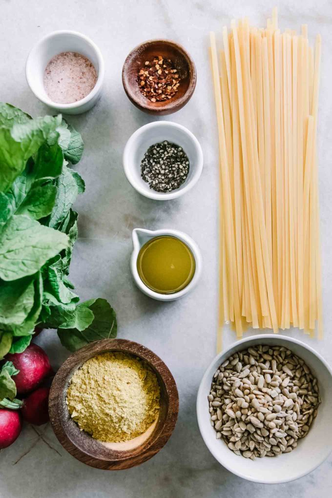 dry pasta, a bunch of radishes, and bowls of nuts, cheese, olive oil, salt and pepper on a white table