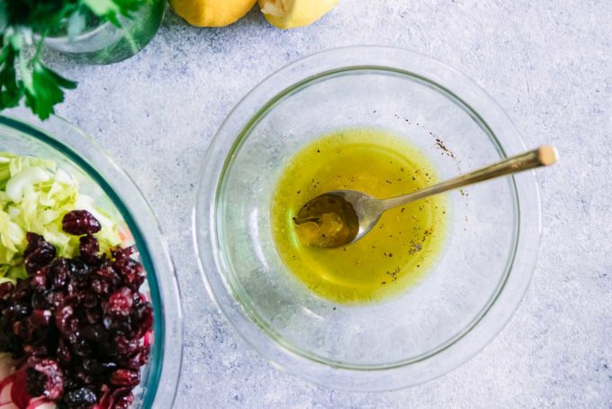 a glass bowl with vinaigrette dressing and a gold spoon on a blue table next to a chopped salad