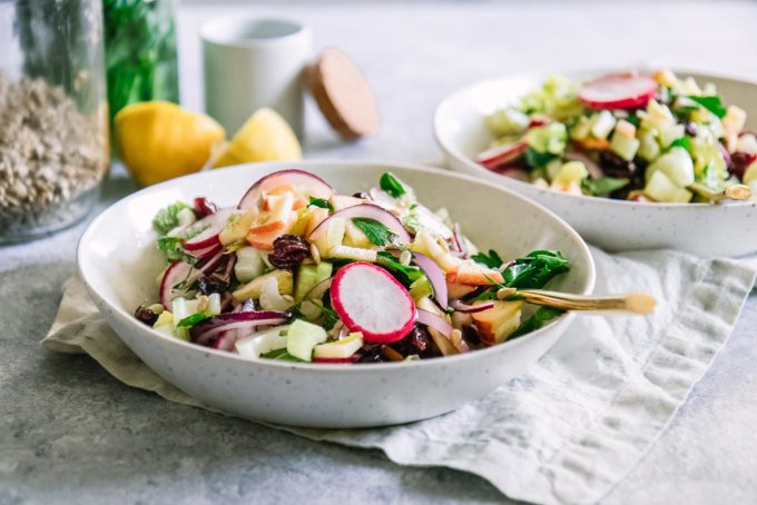 a salad with celery, radishes, onions, apples, and herbs in a white bowl on a white table with a gold fork