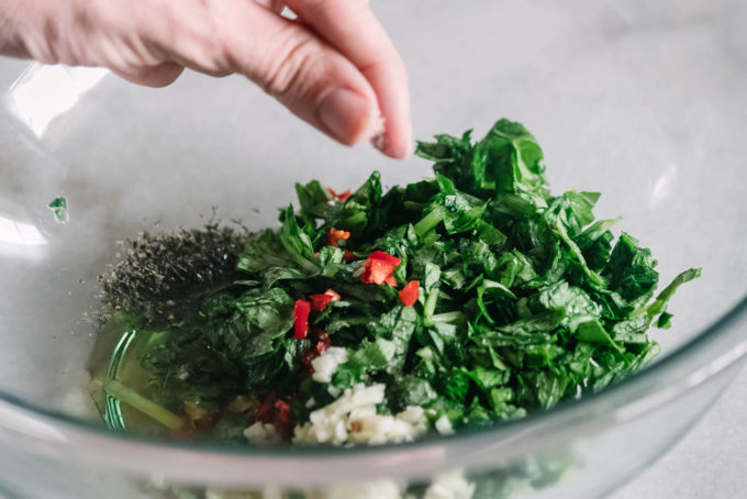 a hand sprinkling salt over chimichurri ingredients in a glass mixing bowl