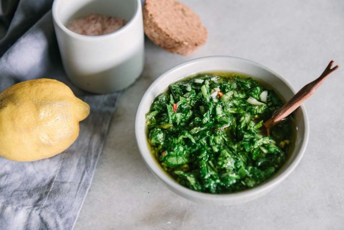 a bowl of chimichurri with a wooden spoon on a white table with radishes and lemons