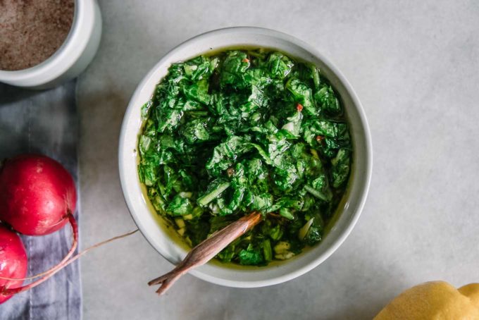 a bowl of chimichurri on a white table with radishes and lemons