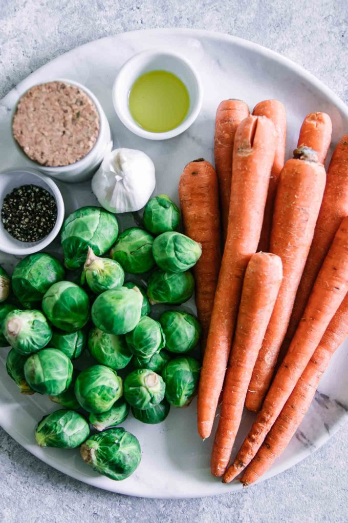 carrots, brussels sprouts, garlic, and bowls of oil and salt on a white cutting board