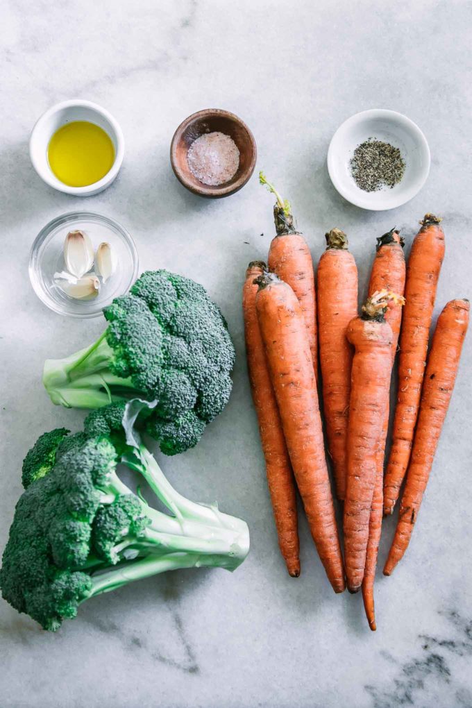 carrots, a head of broccoli, and bowls of garlic, olive oil, salt, and pepper on a marble countertop