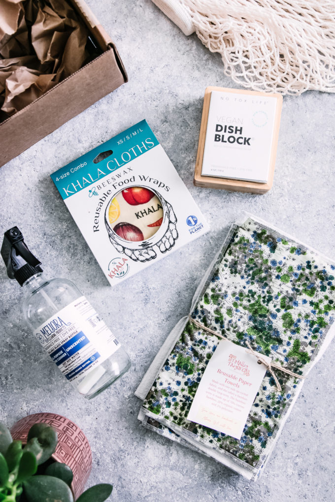 a glass spray bottle, reusable paper towels, bar dish washing soap, and beeswax wrap on a blue table