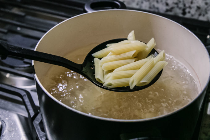a spatula with penne noodles above a pot of pasta boiling in hot water on a stove