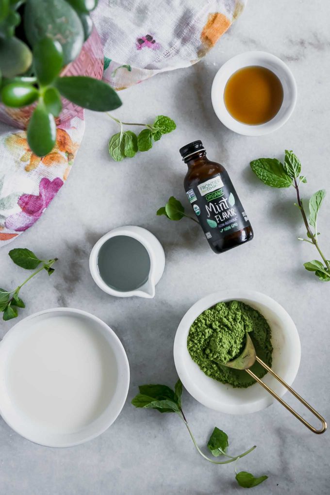 bowls of matcha green tea powder, mint extract, plant-based milk, water, and maple syrup on a white marble countertop with fresh mint