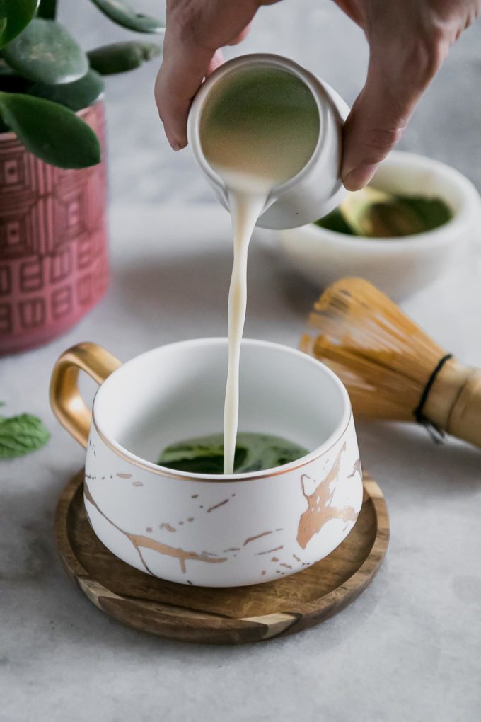 a hand pouring milk into a white ceramic mug filled with matcha green tea