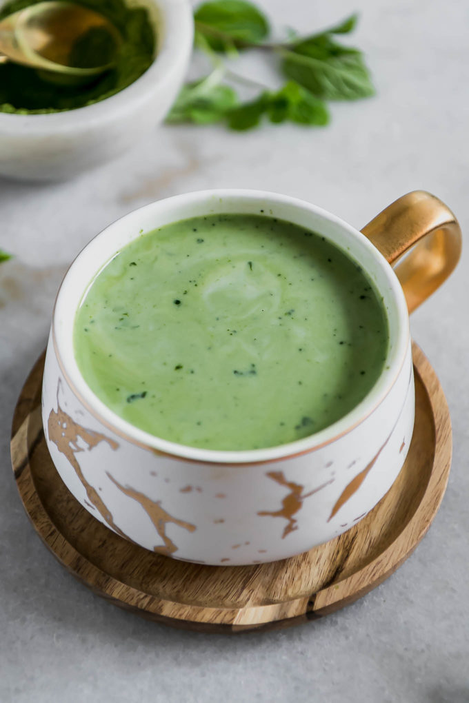 a cup of matcha green tea mixed with milk on a wooden saucer on a white table with mint leaves