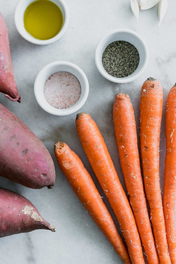 sweet potatoes, carrots, and bowls of salt, pepper, and olive oil on a white marble countertop