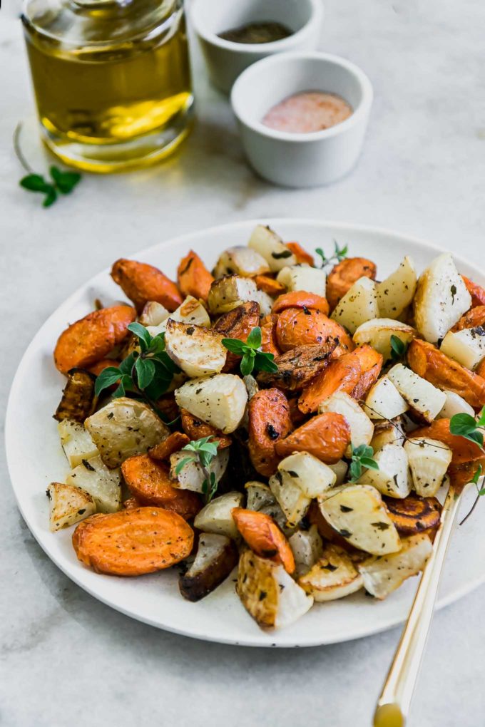 roasted turnips and carrots on a white plate on a white table with a bottle of oil and bowls of salt and pepper in the background