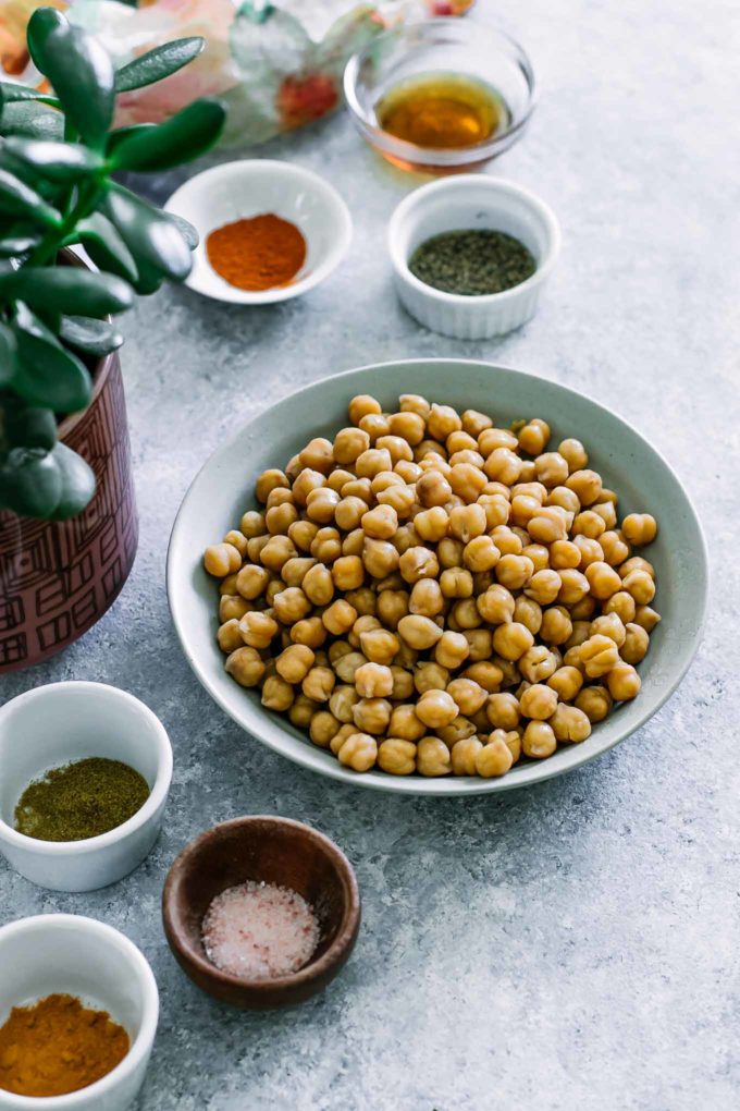 bowls of chickpeas, maple syrup, salt, and other spices on a blue table with a green plant