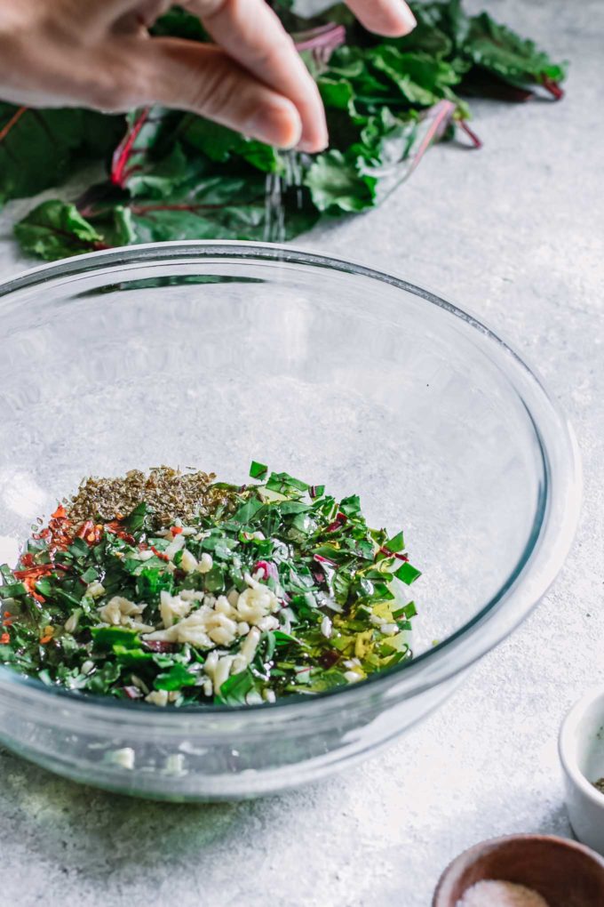 a hand sprinkling salt into a bowl of chopped herbs, garlic, red pepper, and oil