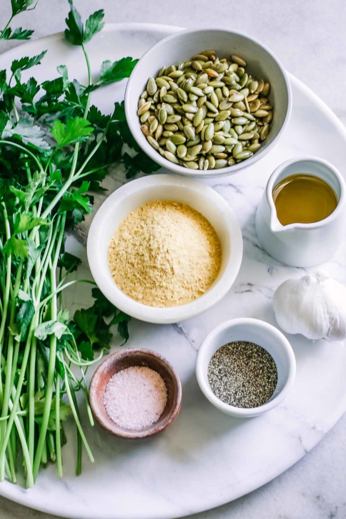 bowls of seeds, nutritional yeast, olive oil, salt, pepper, and a bunch of parsley on a white countertop