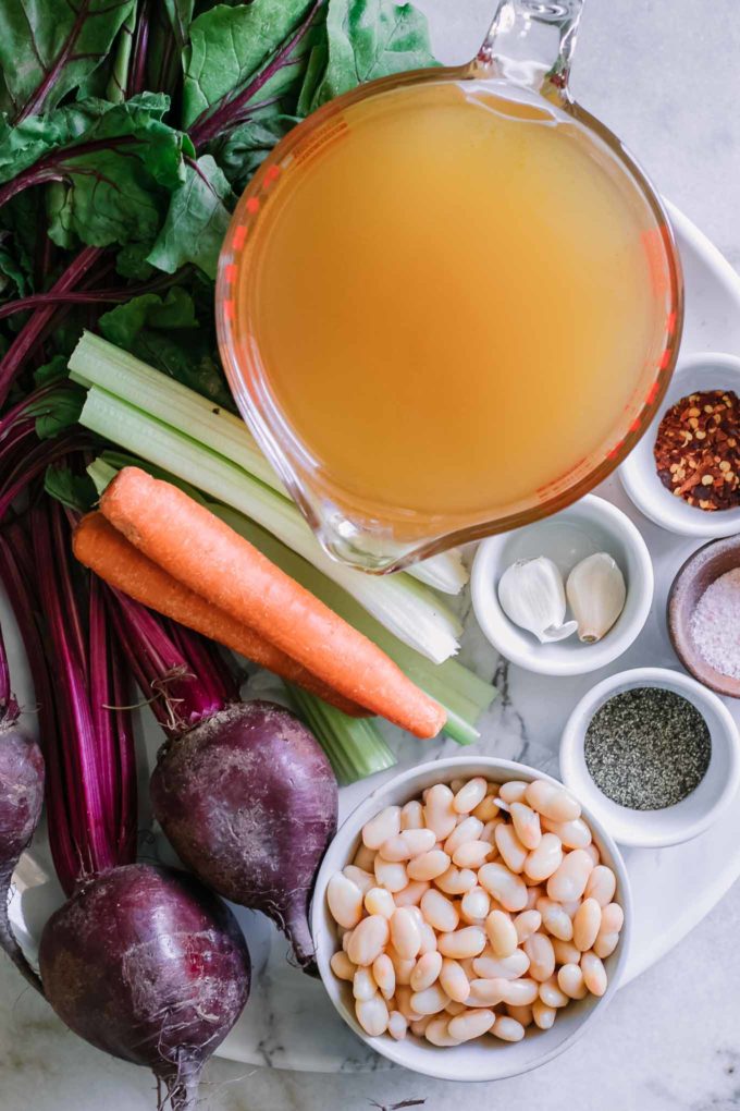 beets, carrots, celery and bowls of white beans, garlic, and vegetable broth on a white countertop