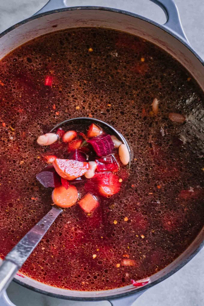 a ladle mixing red beet soup with vegetables inside a soup pot