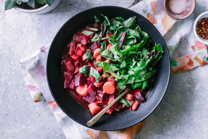 a black bowl of red beet soup with chopped beet greens on a blue table