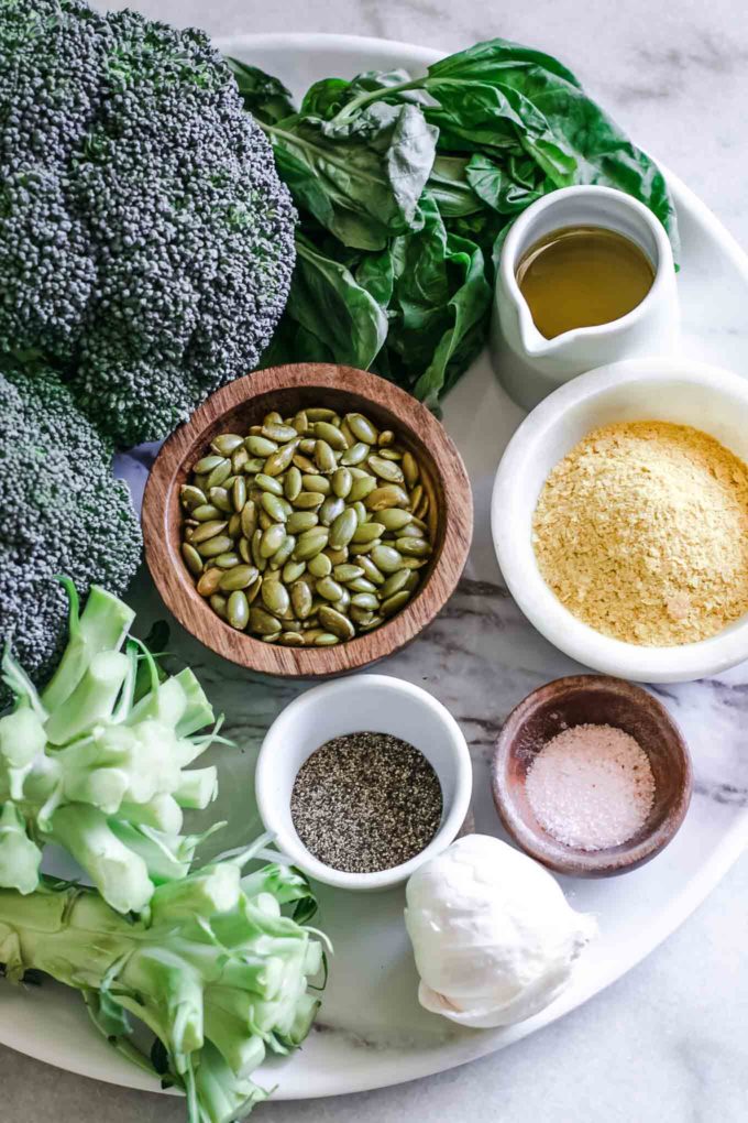 broccoli stems, basil leaves, and bowls of nuts, cheese, oil, salt, pepper, and garlic on a white table