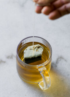 a hand pulling a tea bag out of a glass mug of brewed tea