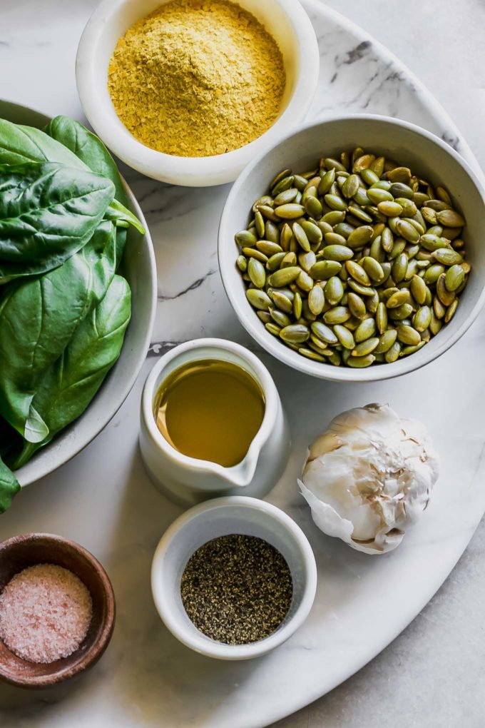 bowls of basil, pumpkin seeds, nutritional yeast, olive oil, salt, pepper, and a whole garlic clove on a white table