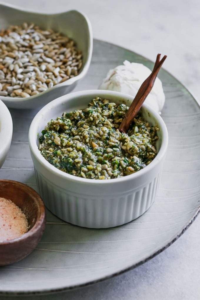 a bowl of sunflower seed pesto with a wooden spoon on a white table with sunflower seeds and salt