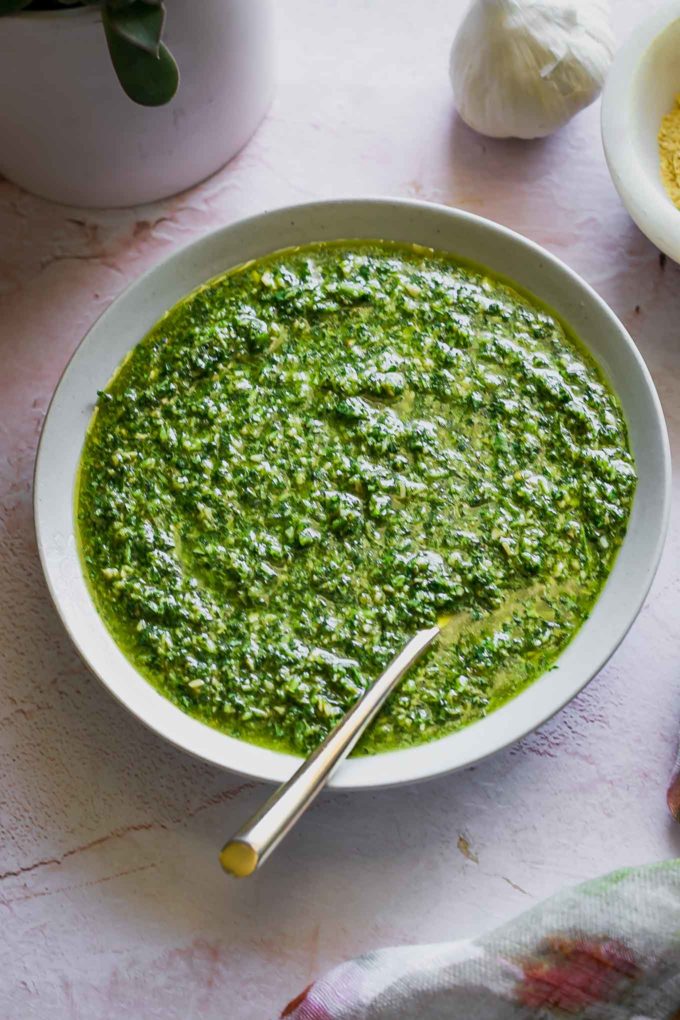 a bowl of pesto with cilantro on a pink table with a bowl of nutritional yeast and a head of garlic