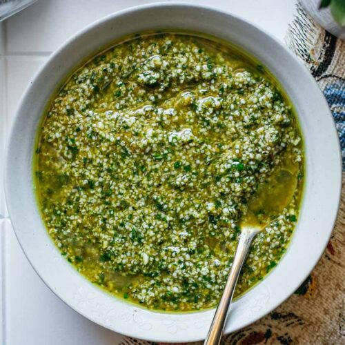 hemp seed pesto in a white bowl with a gold spoon on a tiled counter with a floral napkin