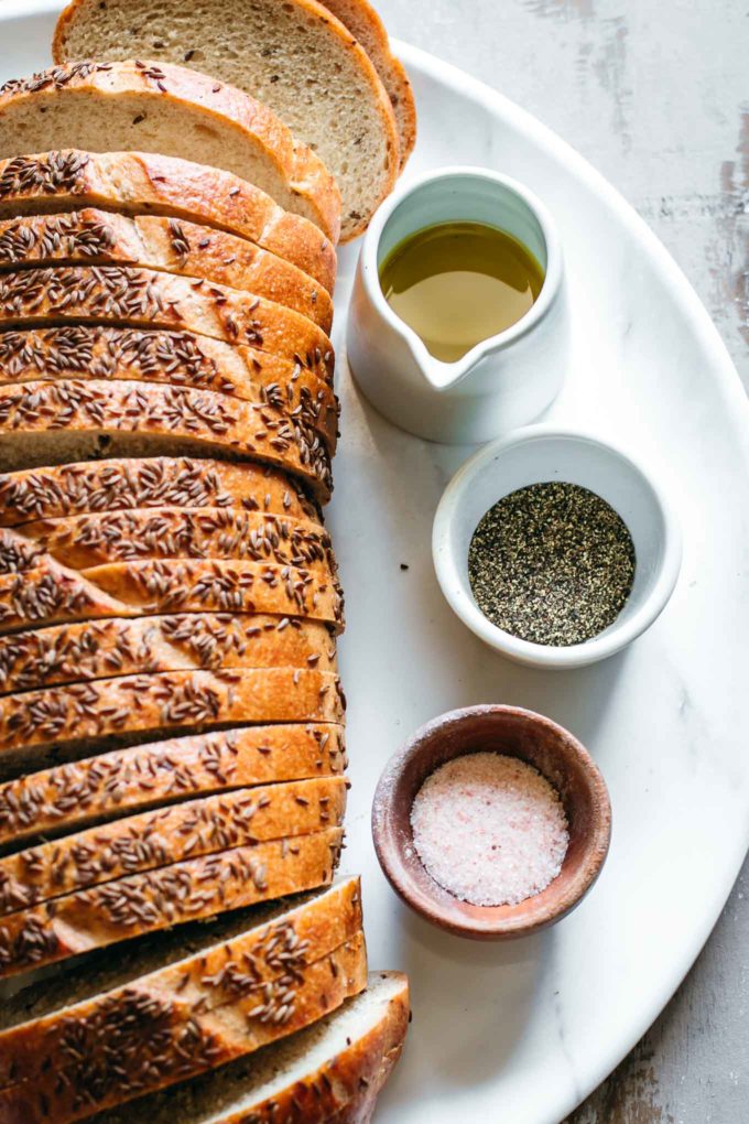 sliced loaf of rye bread with bowls of olive oil, salt, and pepper on a white platter
