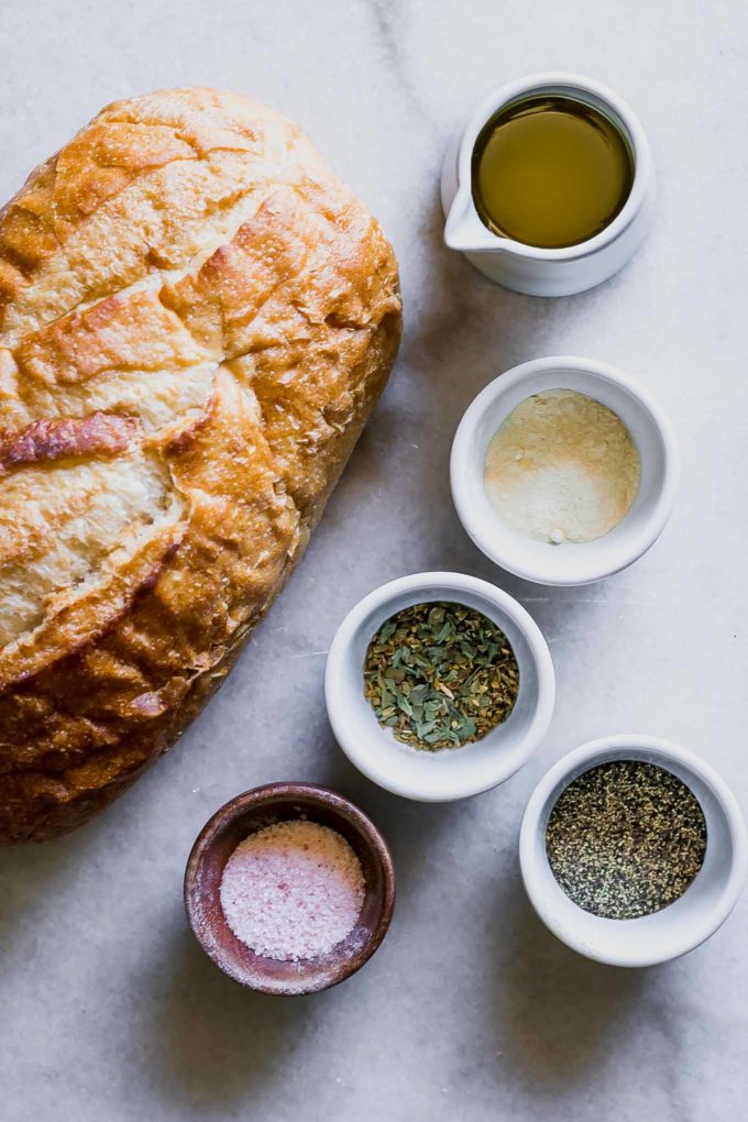 loaf of sourdough bread with bowls of olive oil, salt, pepper and spices on a white counter