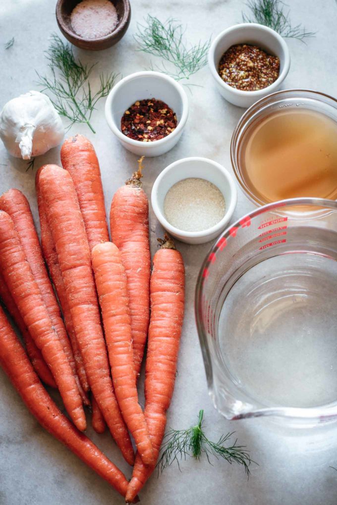 whole carrots and bowls of garlic, mustard seeds, red pepper flakes, salt, sugar, water and vinegar