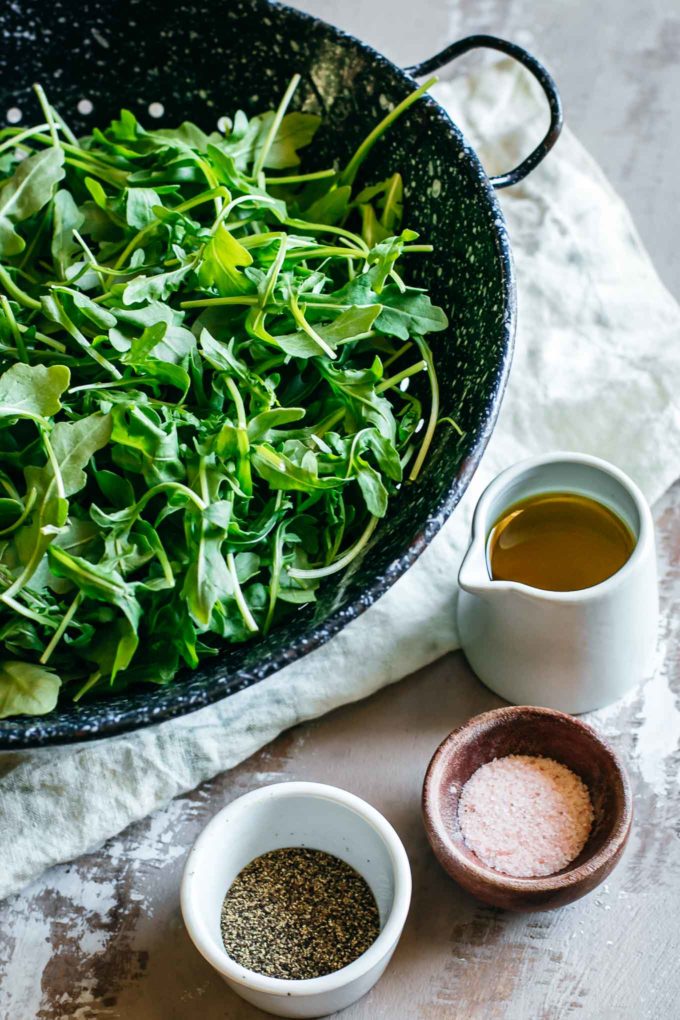 arugula leaves in a colander with bowls of oil, salt, and pepper on a brown table