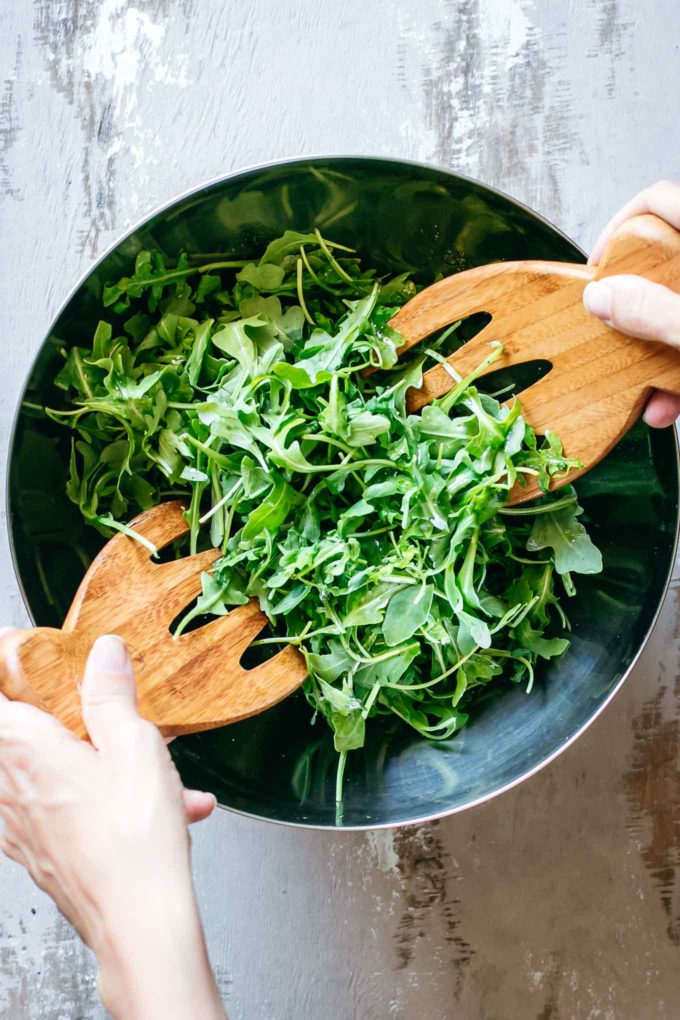 two hands tossing arugula with wooden spoons in a silver mixing bowl