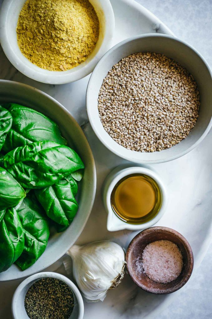 bowls of basil, cheese, sesame seeds, oil, salt, pepper, and garlic on a white table